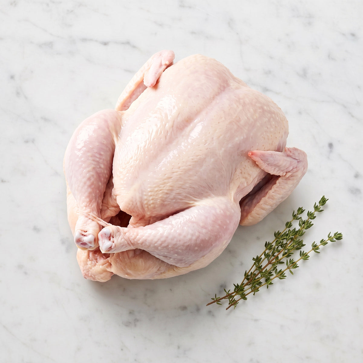Whole raw chicken with trussed legs beside thyme sprigs on white marble
