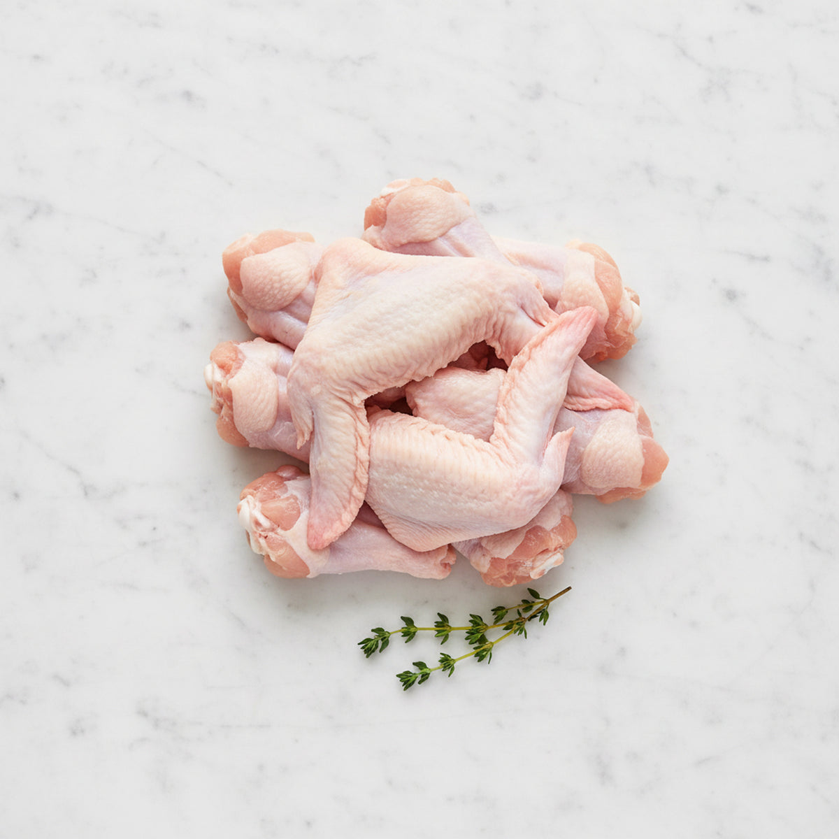 Pile of raw whole chicken wings with a thyme sprig on white marble
