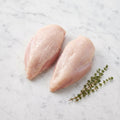 Two raw bone-in, skin-on chicken breasts without wings, with thyme on white marble