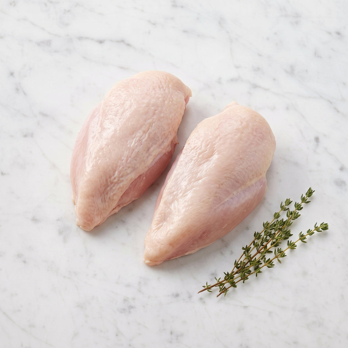 Two raw bone-in, skin-on chicken breasts without wings, with thyme on white marble