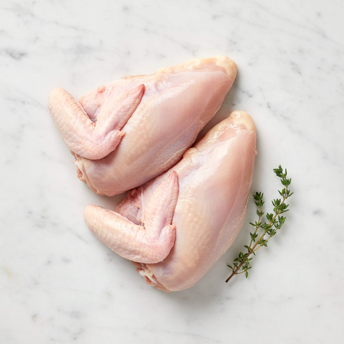 Two raw bone-in chicken breasts with wings attached and a thyme sprig on white marble