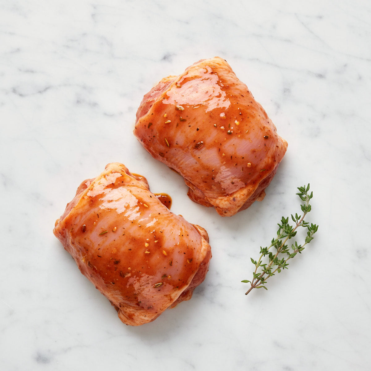 Two chicken pieces in a glossy spiced marinade with a thyme sprig on white marble