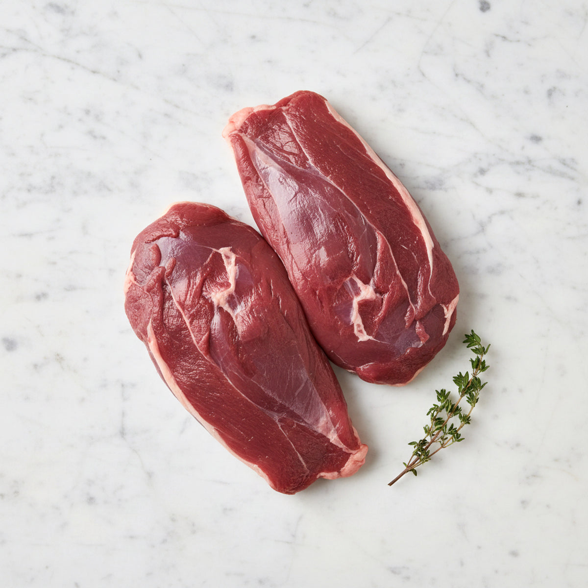 Two raw boneless duck breasts, meat side up, with thyme on white marble