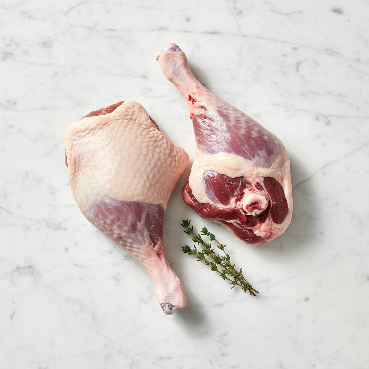 Two raw duck legs, one skin side up and one meat side up, with thyme on white marble
