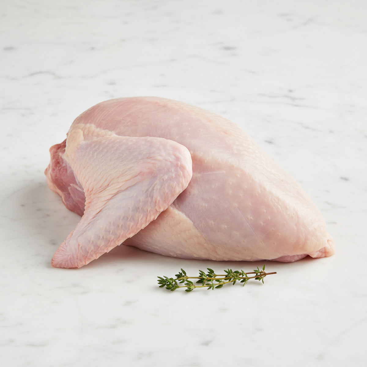 Raw bone-in turkey breast with the wing attached and a thyme sprig on white marble