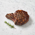 A Grilled Beef Rib Steak, bone-in with char marks and cracked pepper, rosemary alongside, on marble