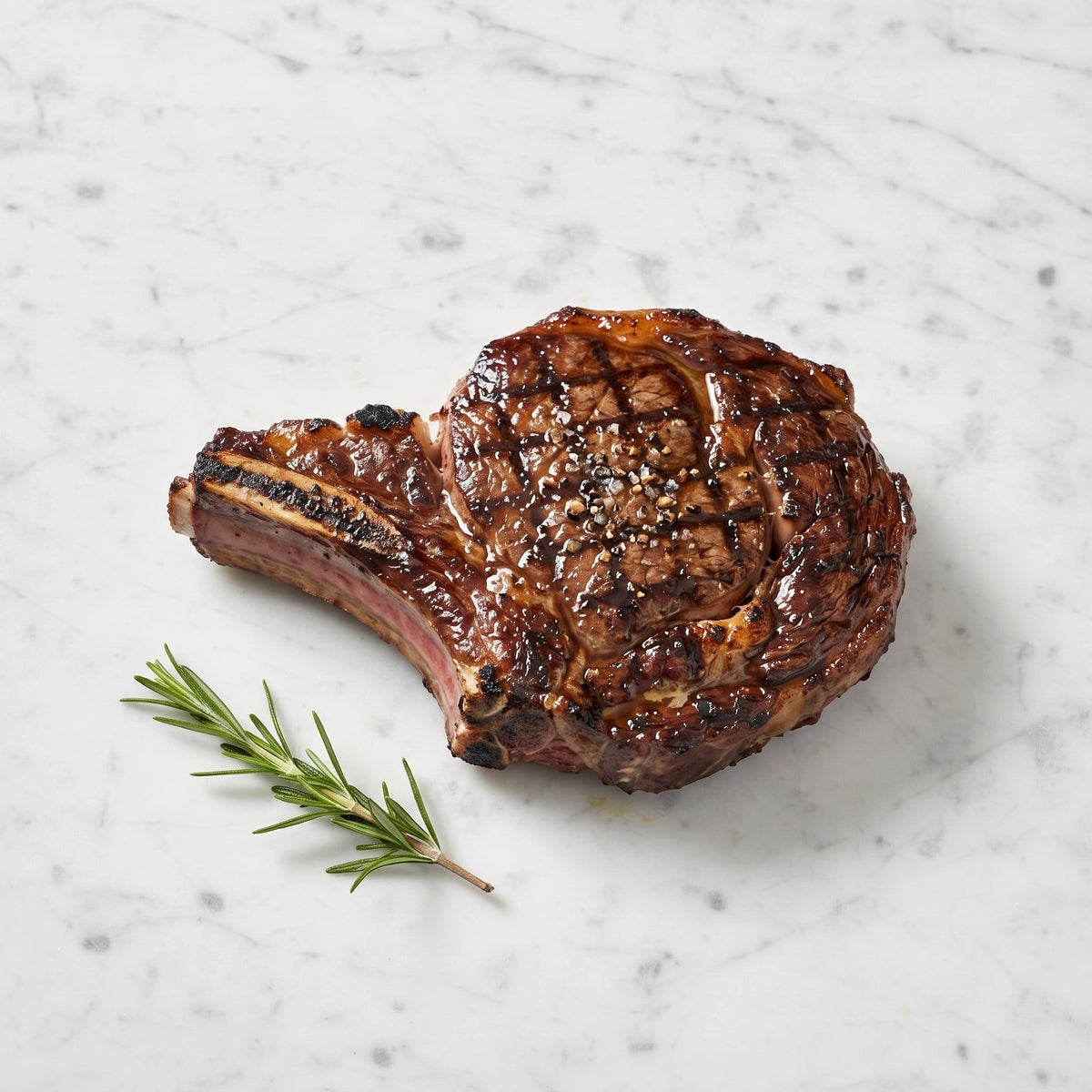 A Grilled Beef Rib Steak, bone-in with char marks and cracked pepper, rosemary alongside, on marble