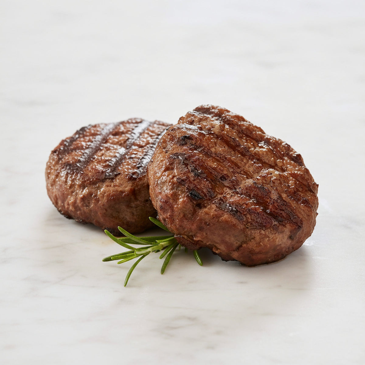 Two TK Char Broiled Beef Burgers with grill marks and a sprig of rosemary on a marble surface