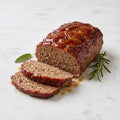 TK Meatloaf with a glazed top, partly sliced, with rosemary and sage on a marble surface
