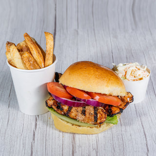 Grilled chicken breast on a bun with tomato, red onion and lettuce, with fries and coleslaw on a wooden table